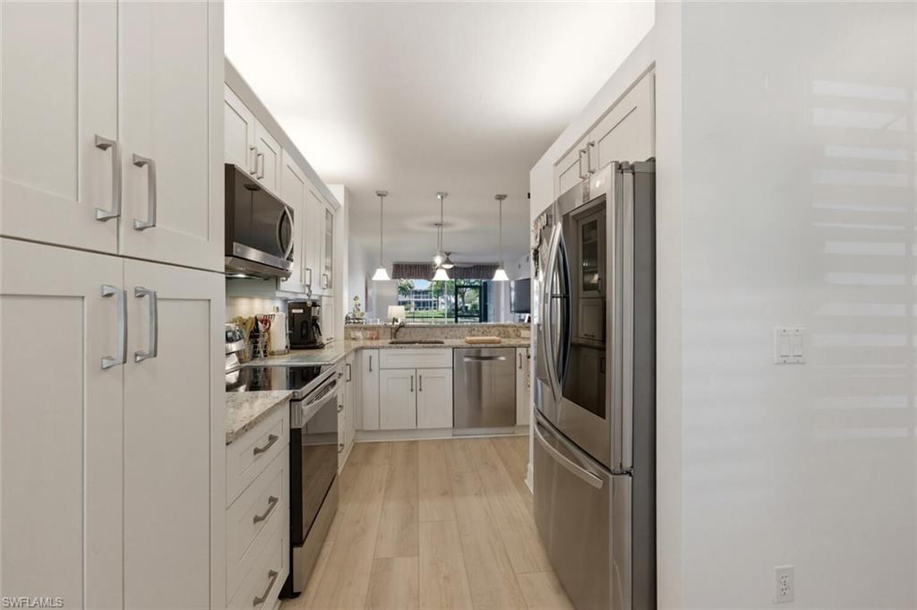 Galley kitchen with white cabinets, stainless steel appliances, stone counters, and light wood flooring.