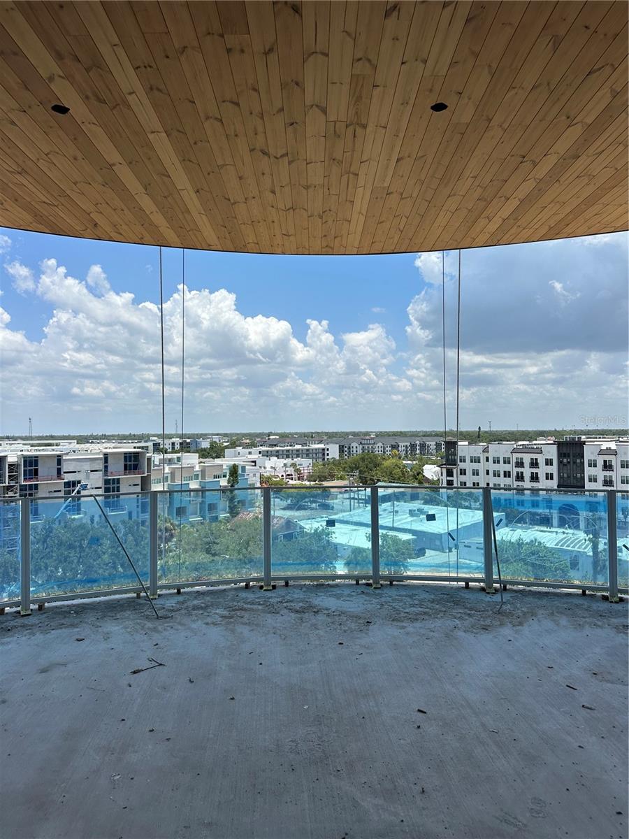 High-rise balcony with a curved glass railing, wood-plank ceiling, and city view.