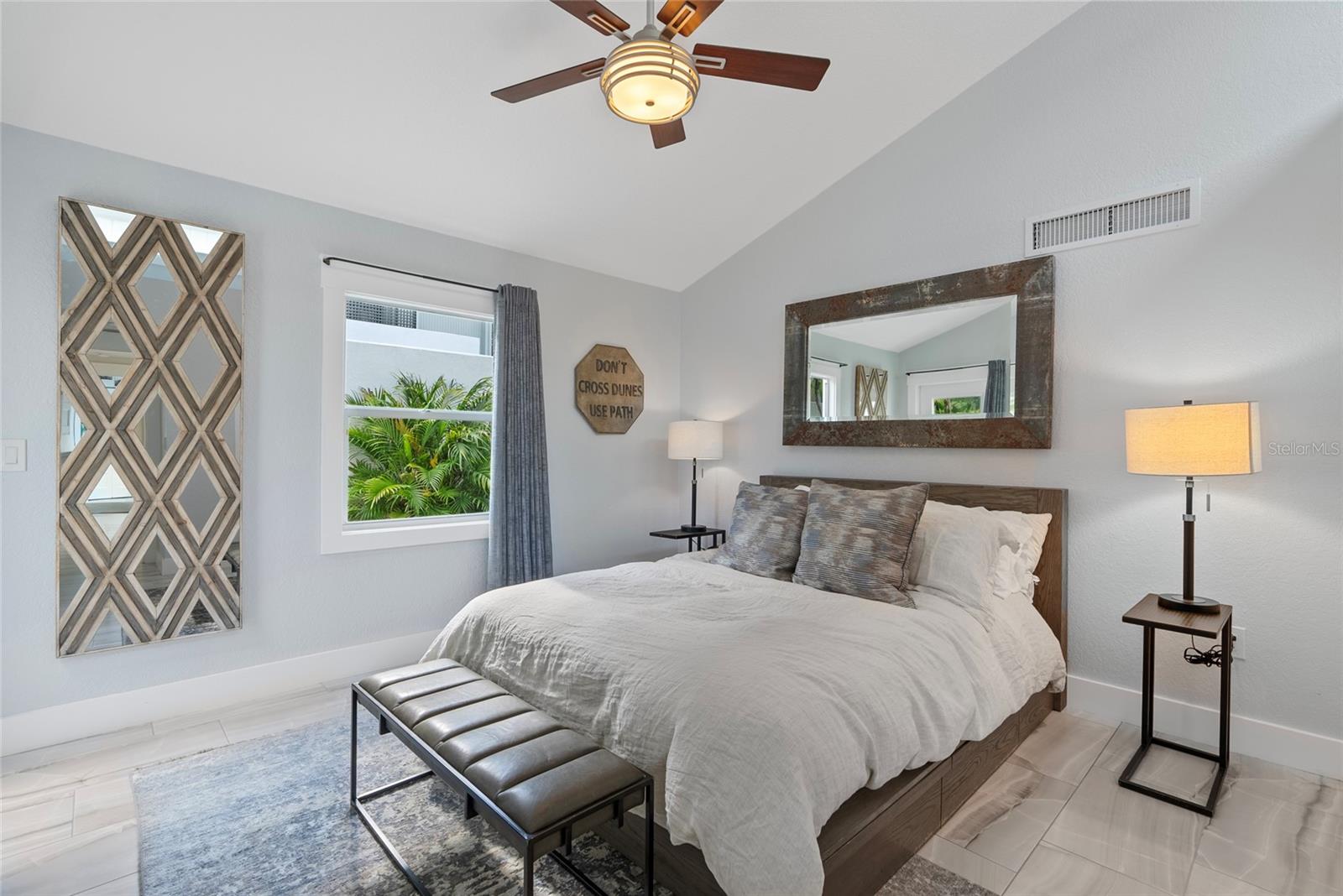 Bedroom with a white bed, dark wood headboard, padded bench, and window overlooking tropical greenery.