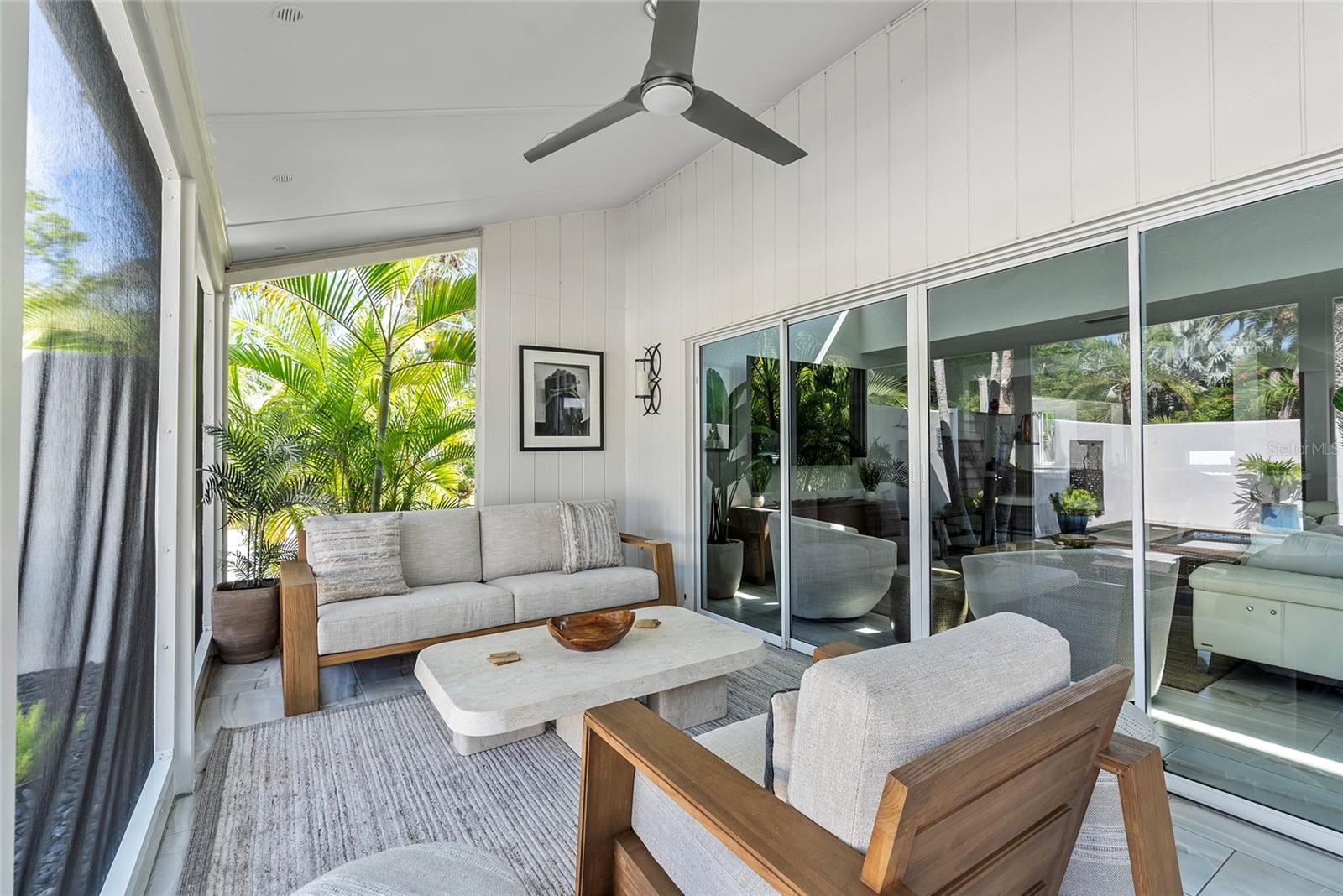 Covered patio with wood-framed sofas and a stone coffee table beside tropical greenery.