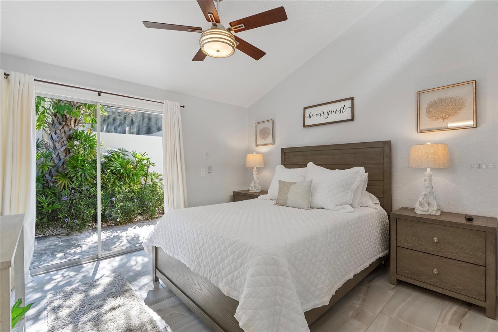 Furnished bedroom with a white bed and sliding glass door overlooking a leafy garden.