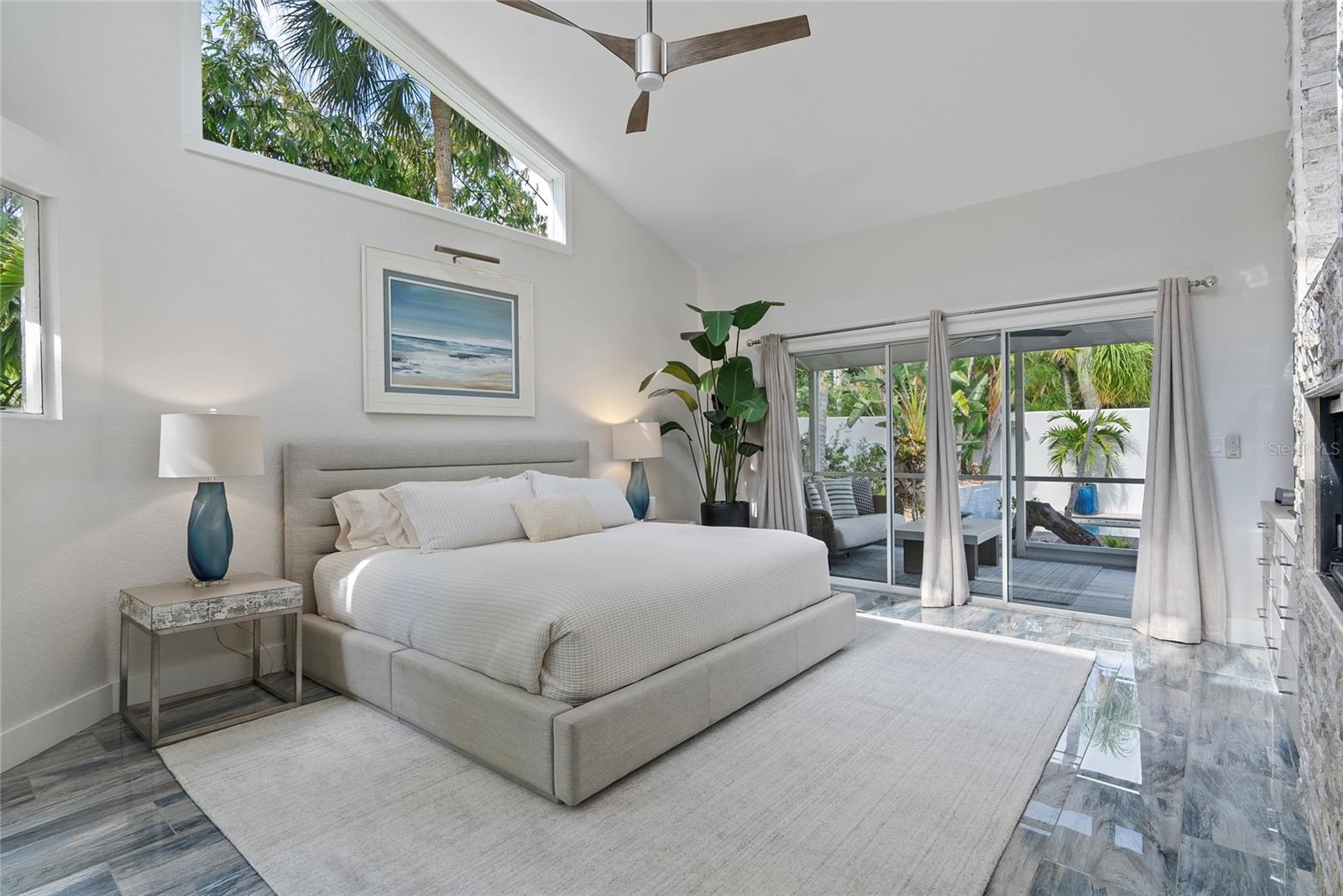 Staged bedroom with a large upholstered bed, vaulted ceiling, and sliding glass doors to a tropical patio.