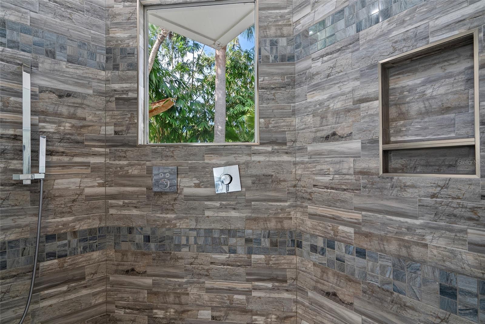 Tiled shower with a chrome fixture, recessed shelves, and a window overlooking tropical greenery.