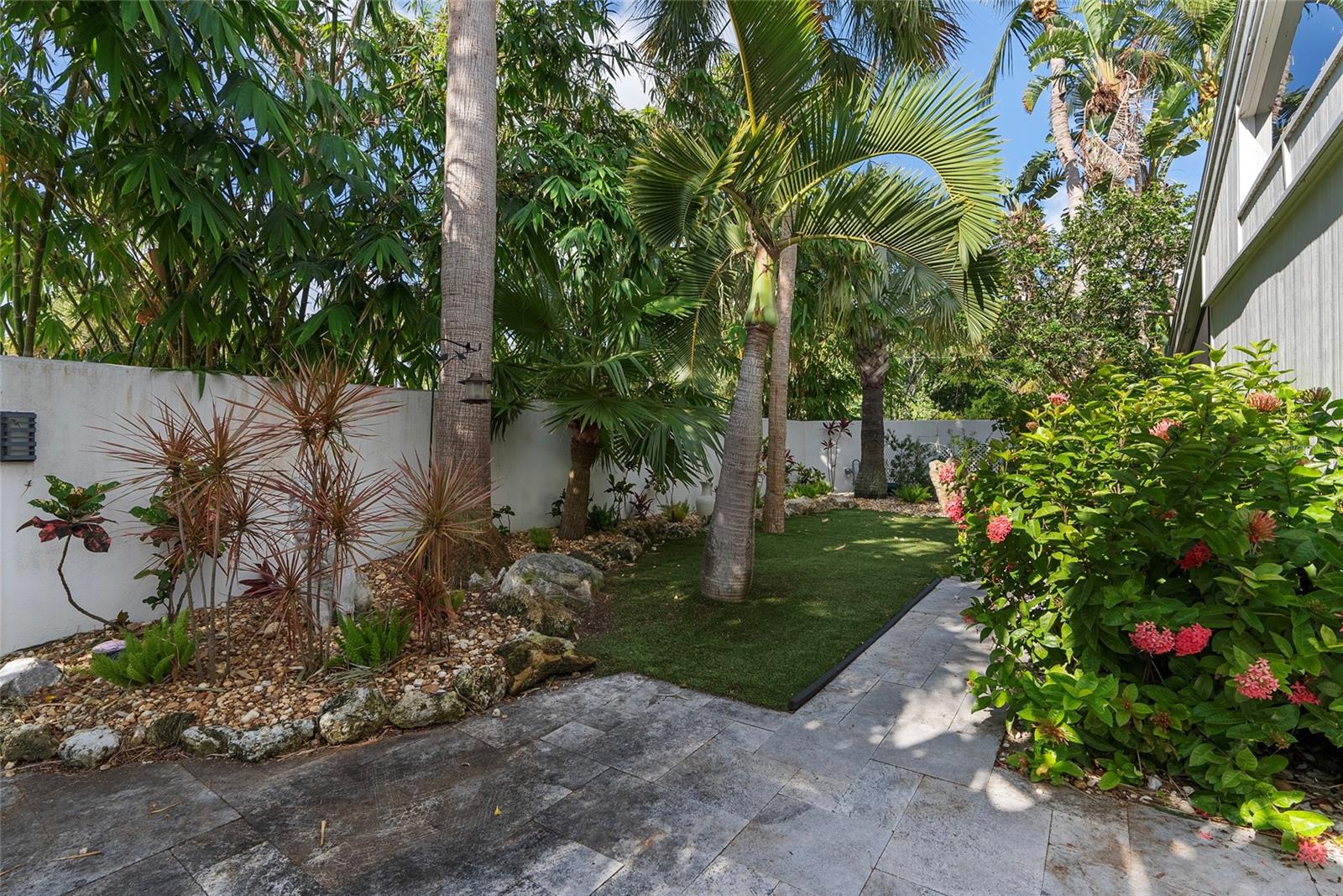 Tropical courtyard with a stone patio, palms, flowering shrubs, and a small lawn.