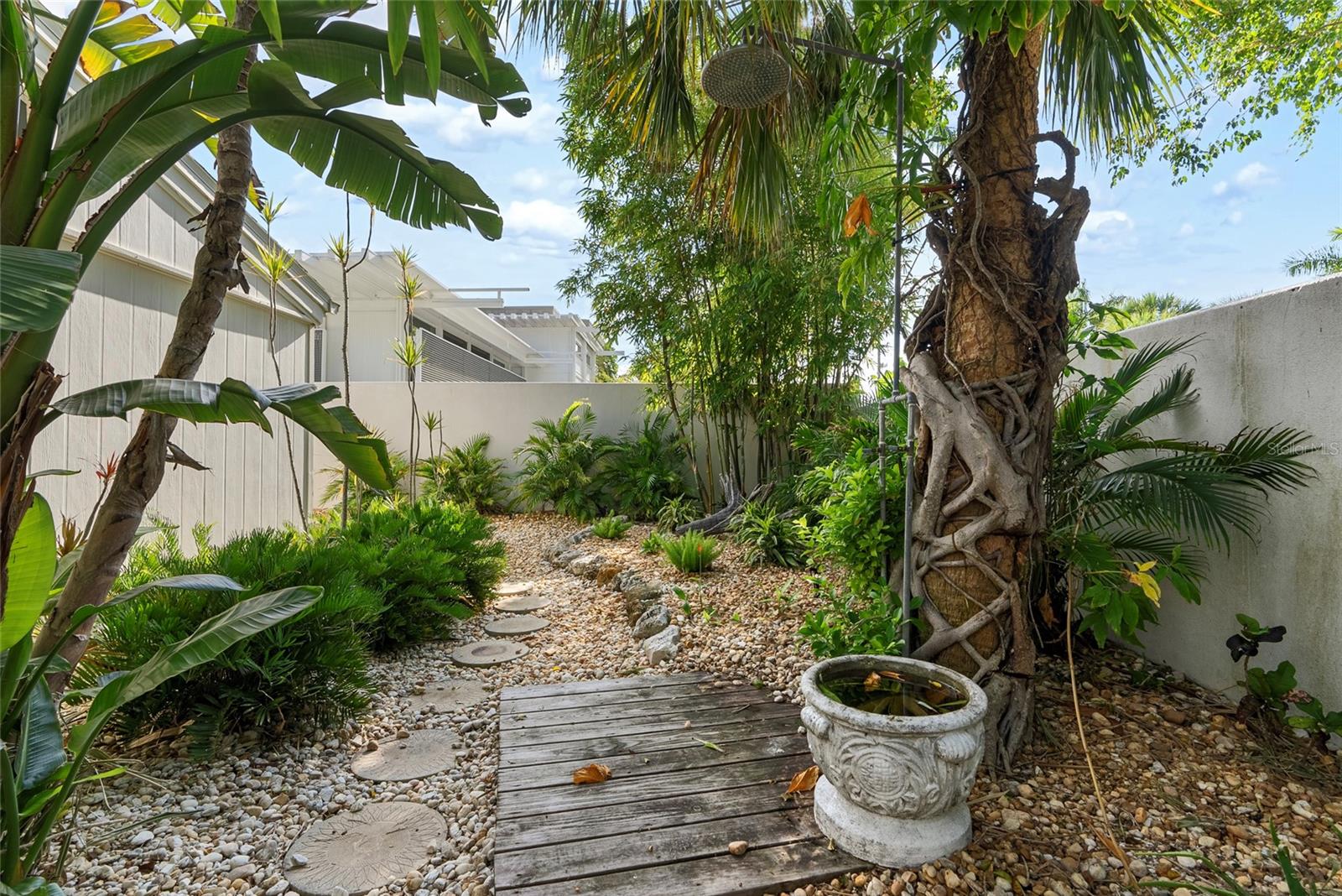 Tropical courtyard garden with a pebble path, wooden walkway, palms, and a carved stone planter.