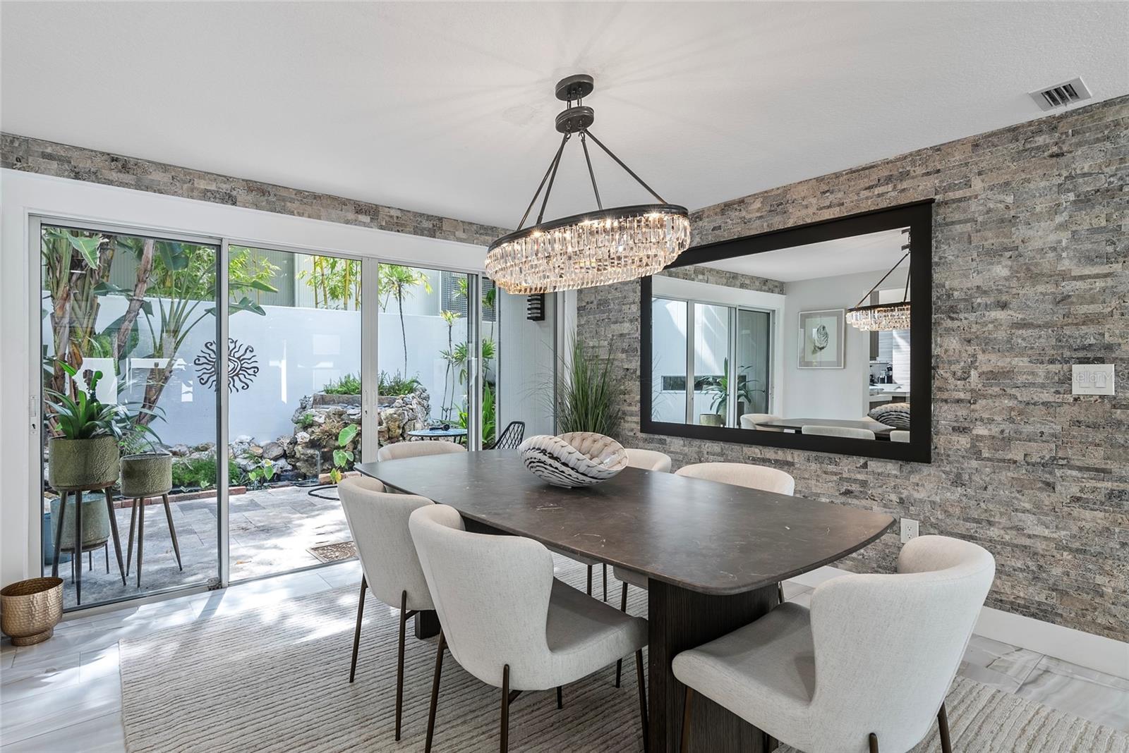 Dining room with a dark table, crystal chandelier, stone walls, and sliding doors to a tropical garden.