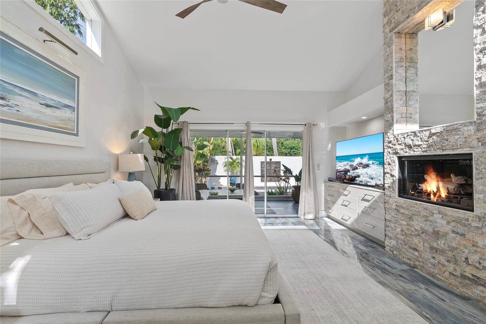 Staged bedroom with a vaulted ceiling, stone fireplace, and glass doors opening to a landscaped patio.