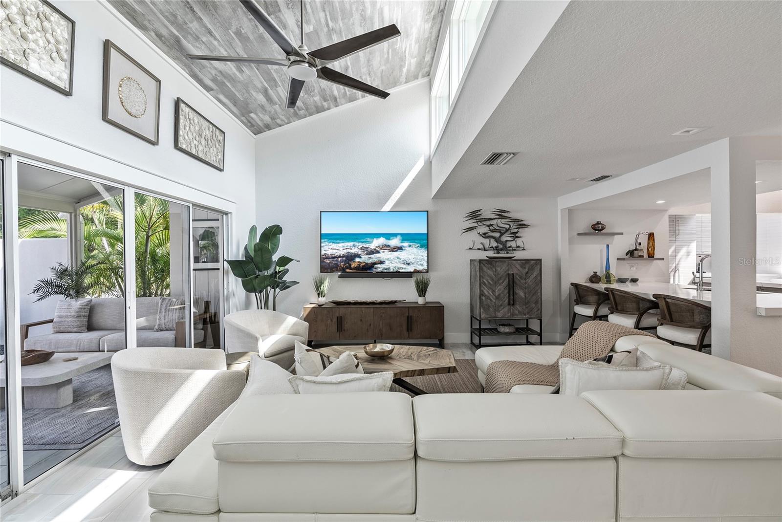 Living room with vaulted ceiling, white seating, and sliding doors to a tropical patio.