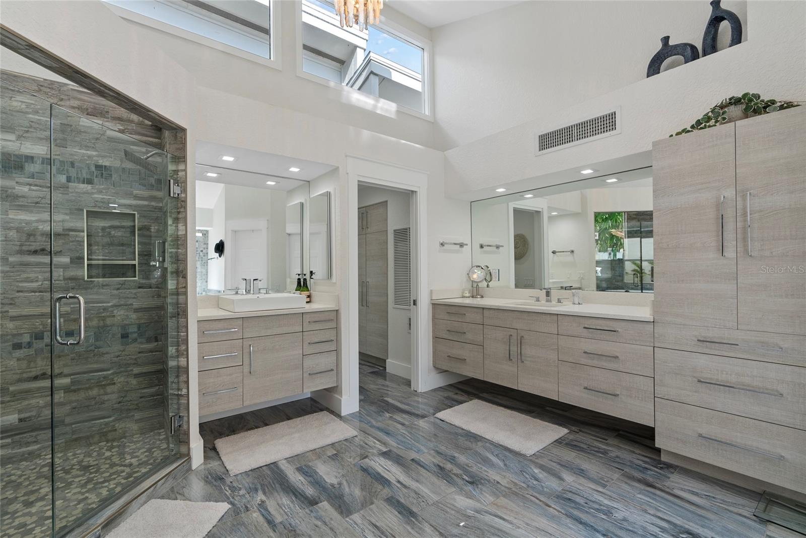Modern bathroom with a glass walk-in shower, double vanity, gray stone tile, and high clerestory windows.