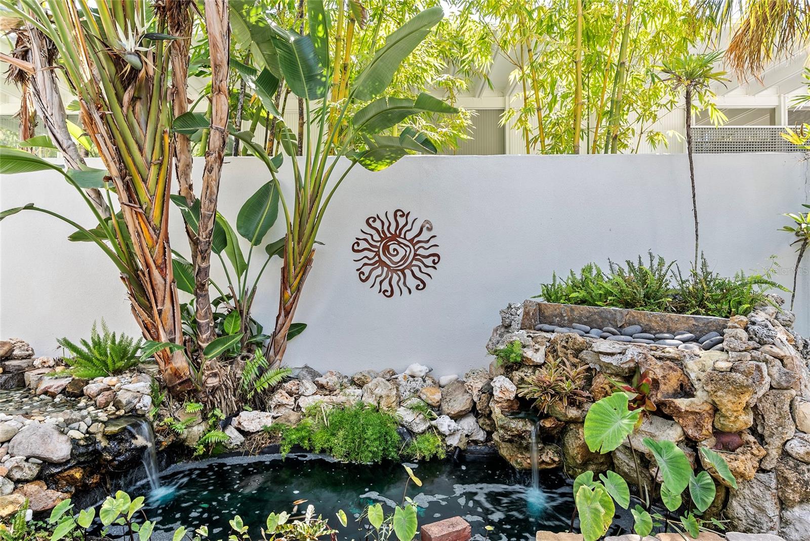 Rock-lined garden pond with bubbling waterfalls, tropical plants, and a sun ornament on a white wall.