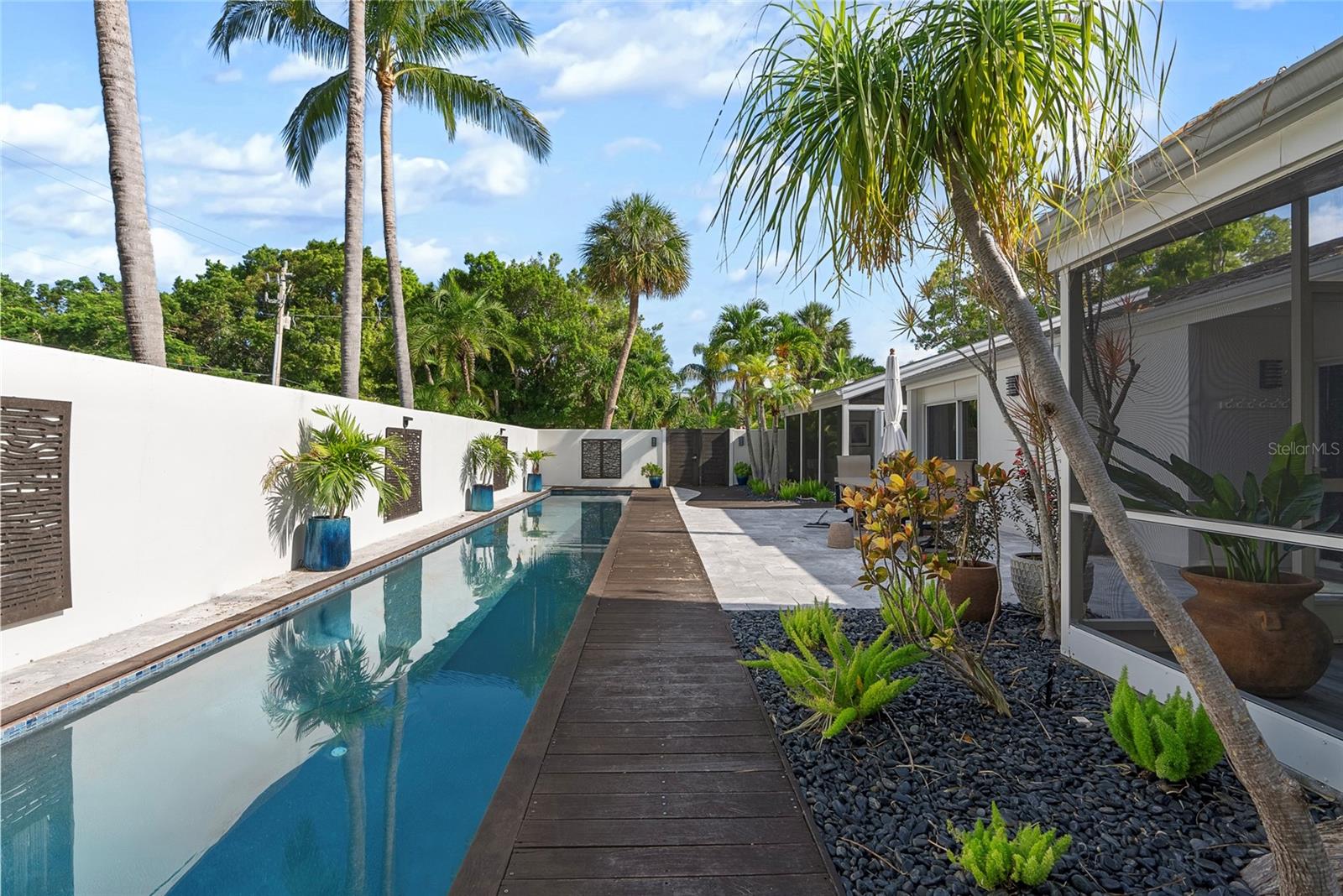 Long courtyard pool beside a modern home, bordered by a dark wood walkway and tropical palms.