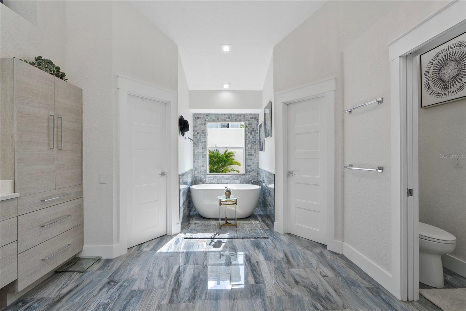 Modern bathroom with a freestanding soaking tub beneath a window and gray tile flooring.