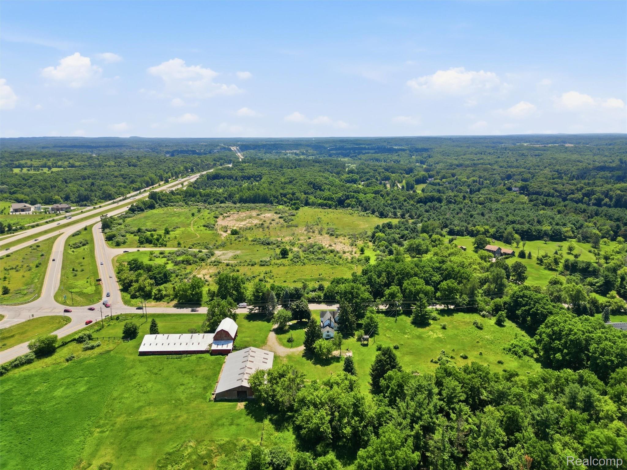 Aerial view of green acreage with a house, barns, wooded areas, and nearby roads.
