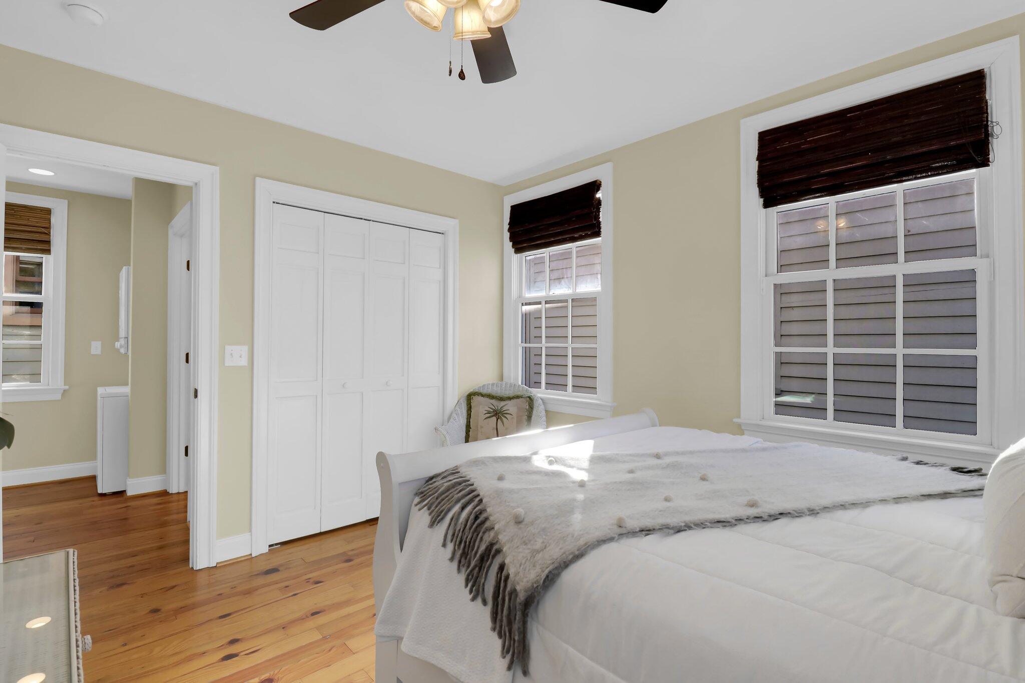 Bedroom with a white bed, hardwood floors, paneled closet doors, and two windows with dark blinds.