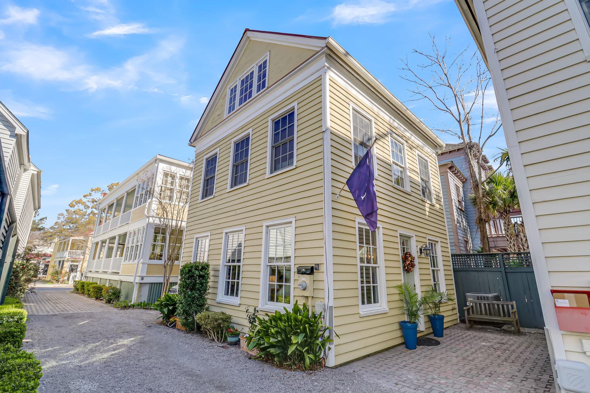Pale yellow multi-story house with white trim, a purple flag, and a brick-paved courtyard.