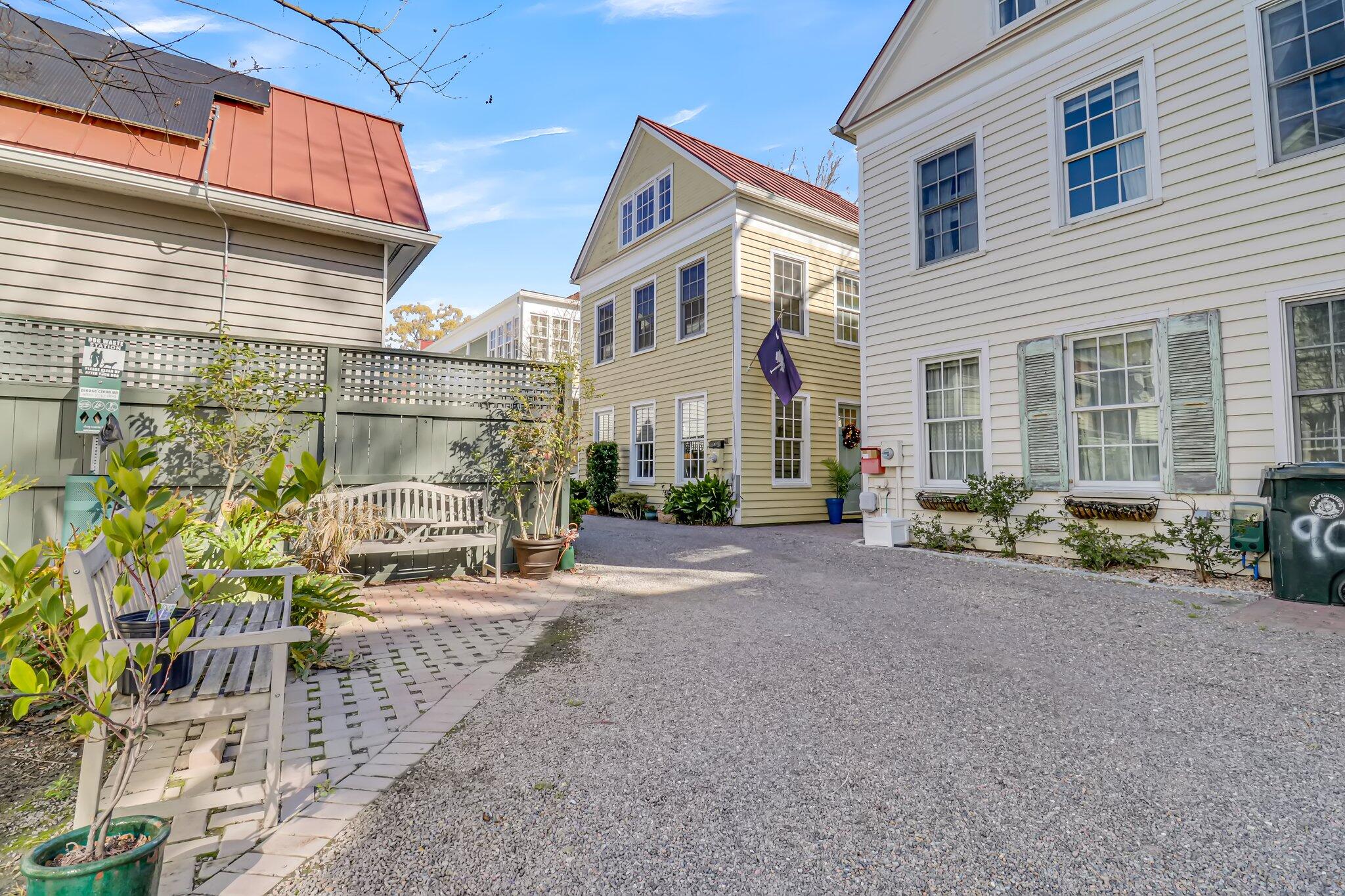 Gravel courtyard between clapboard houses, with a white bench and potted plants along a paved seating area.