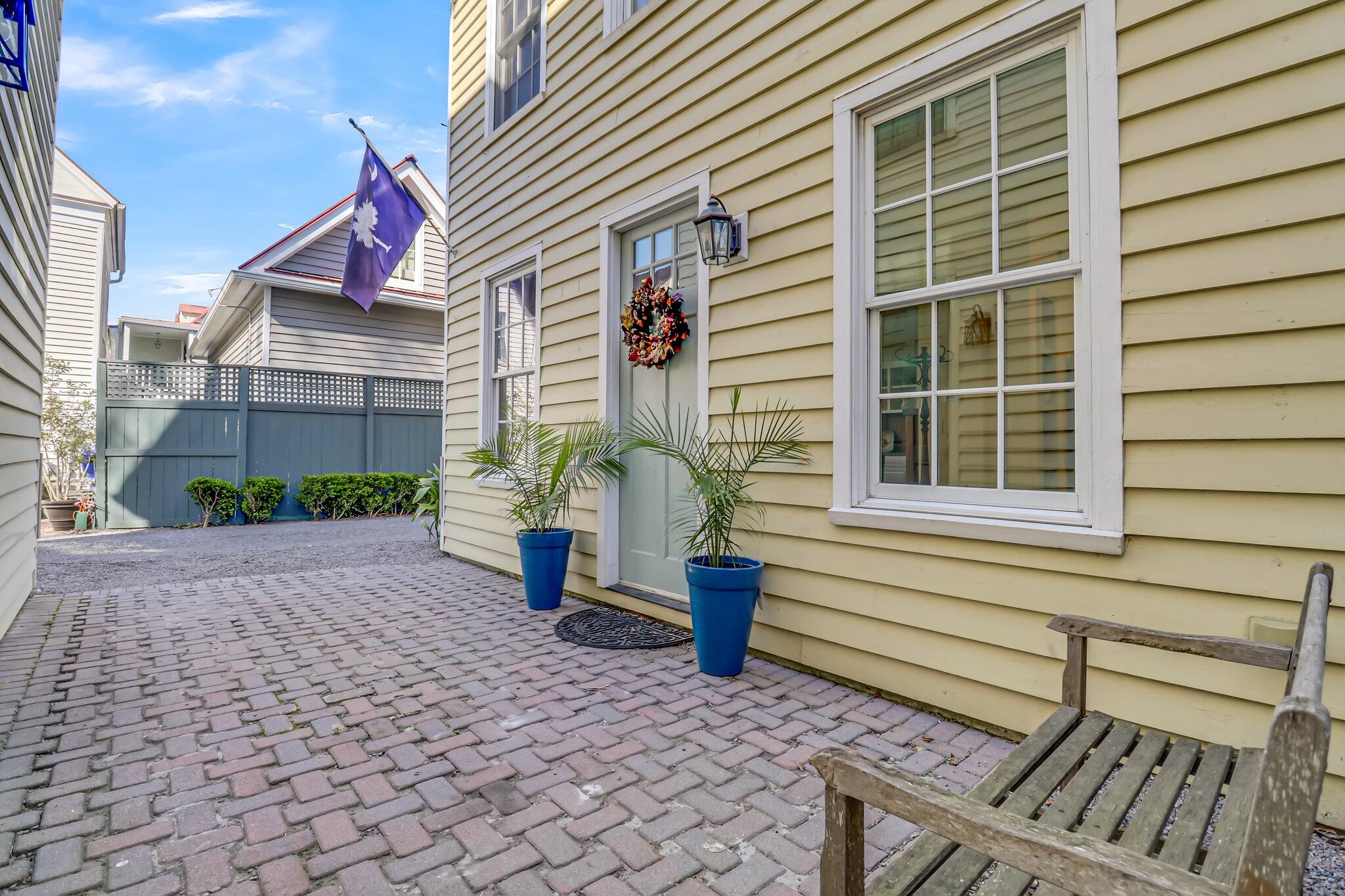 Paved courtyard beside a yellow clapboard home, with a wreath-decorated entry, potted palms, and wooden bench.