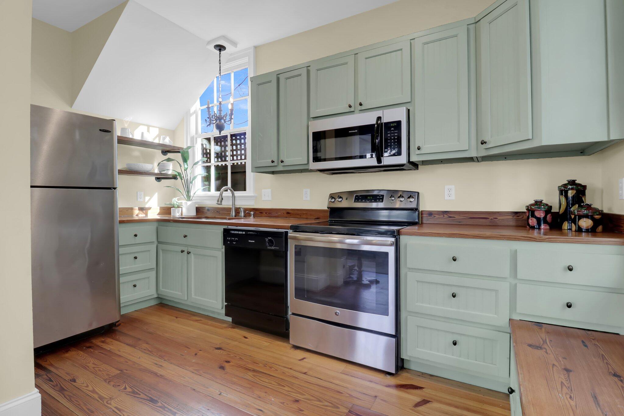 Kitchen with sage-green cabinets, wood counters and floors, stainless appliances, and a tall sunlit window.