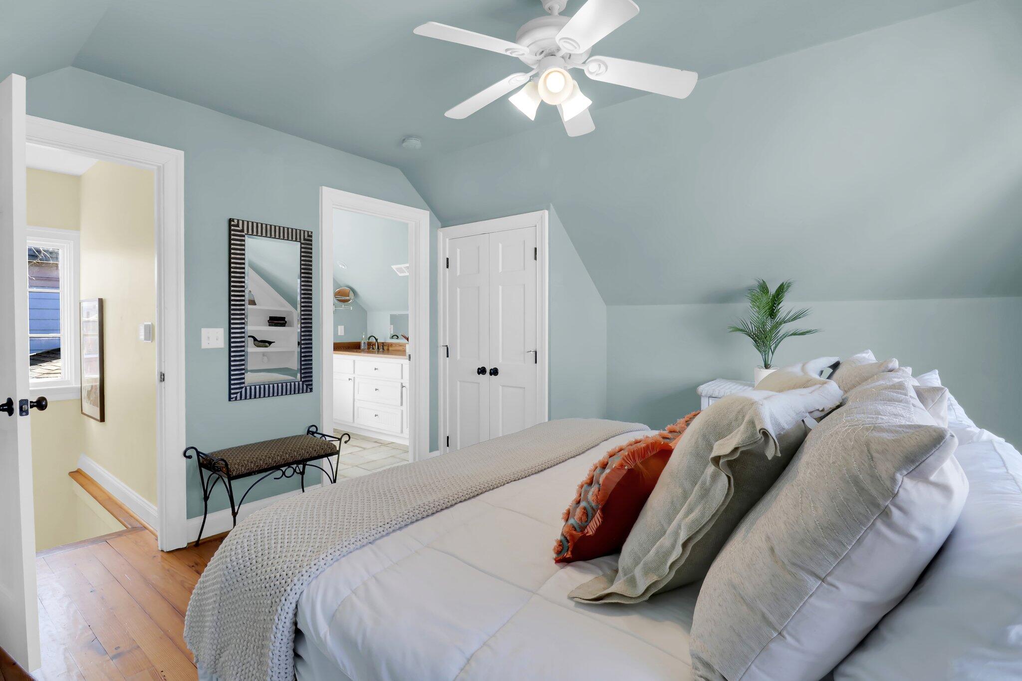 Bedroom with a pale blue sloped ceiling, large bed, ceiling fan, and adjoining bathroom vanity.