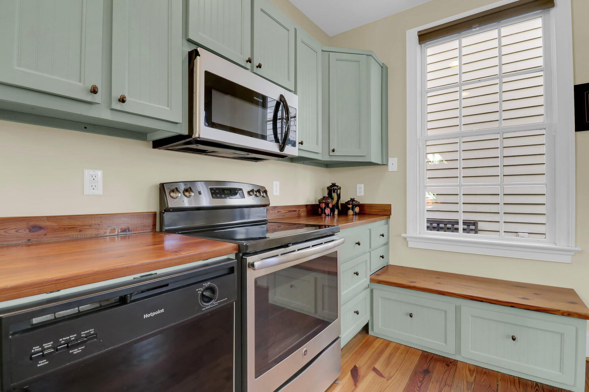 Kitchen with pale green cabinets, wood counters and floors, stainless steel appliances, and a large window.