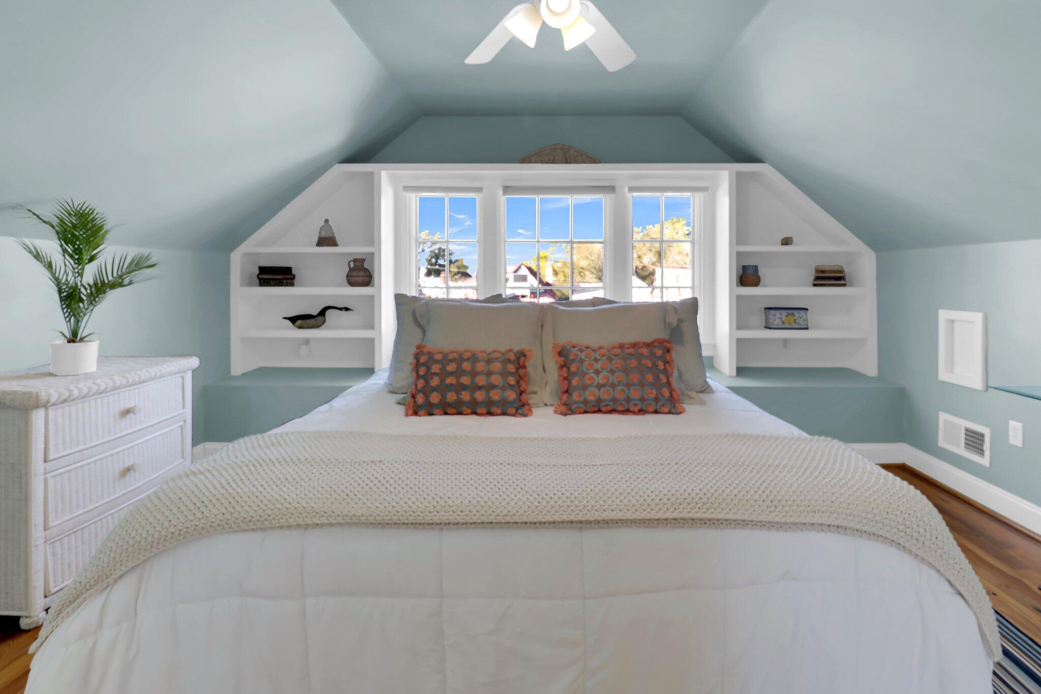 Attic bedroom with a large white bed, built-in shelves, and three windows behind the headboard.