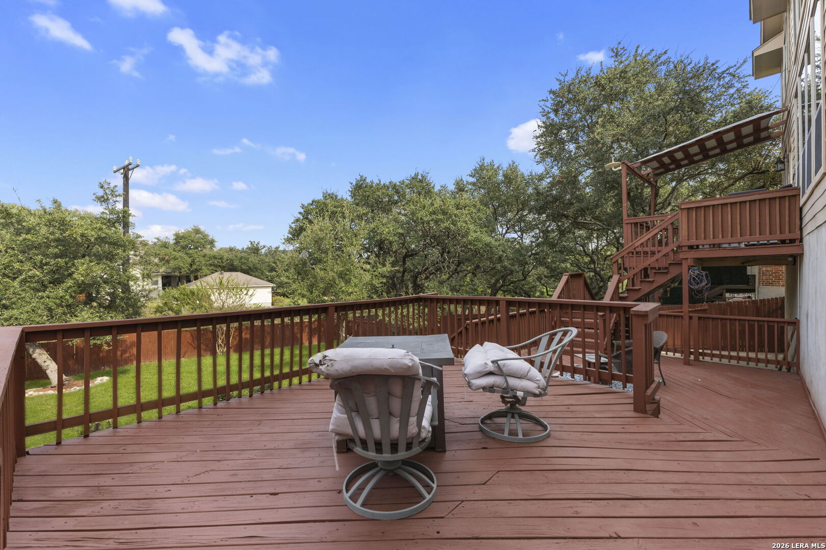 Spacious wooden deck with cushioned chairs, stairs, and a view of mature trees.