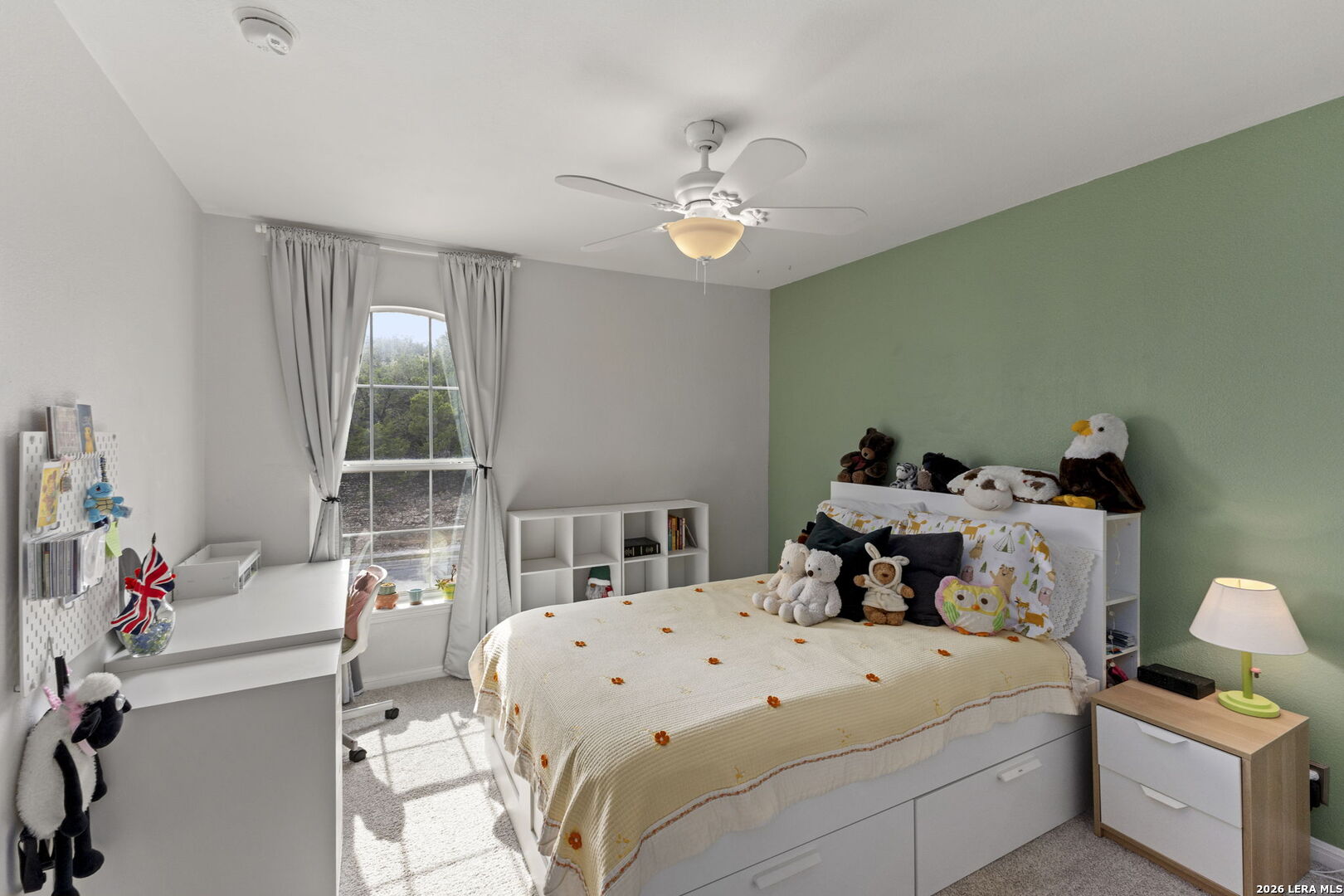 Bedroom with a white bed, plush toys, green accent wall, and desk beside a tall window.
