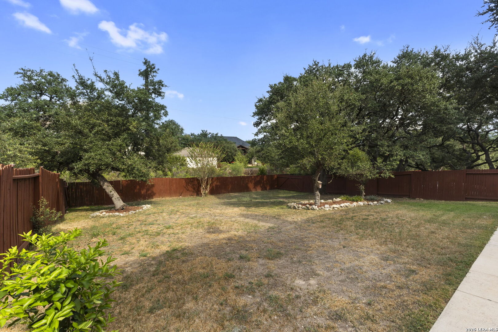 Fenced backyard with mature trees, a patchy lawn, and rock-edged landscaping.