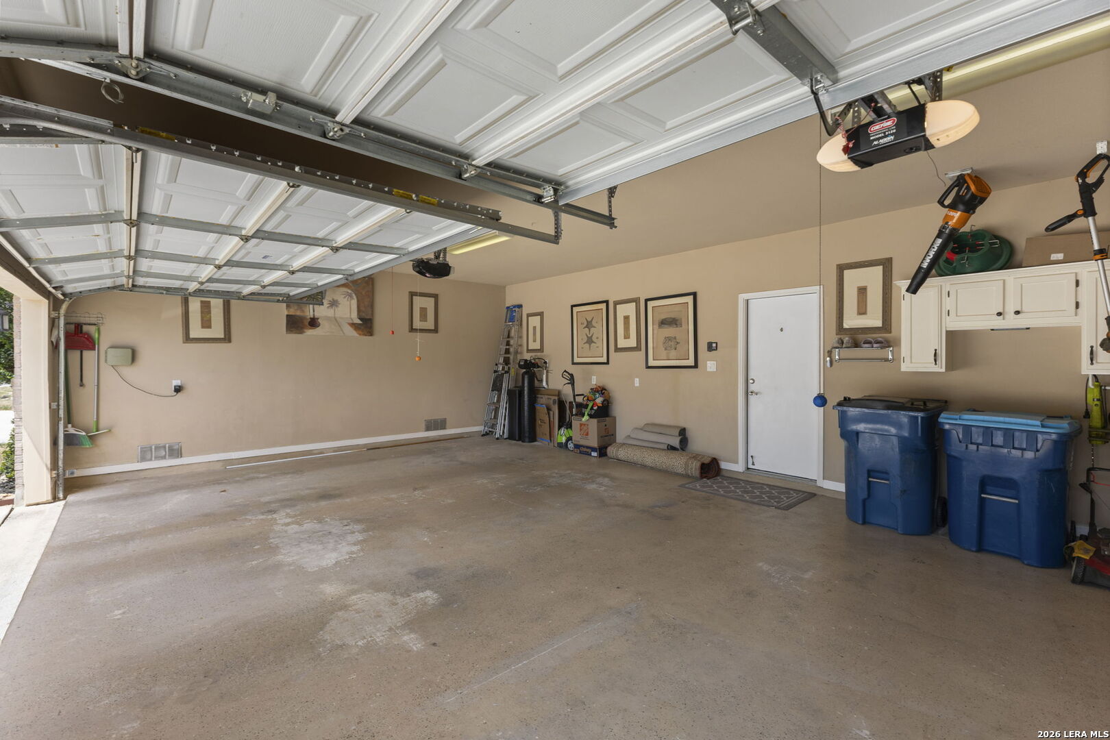 Two-car garage with a raised door, concrete floor, white cabinets, blue bins, and stored tools.