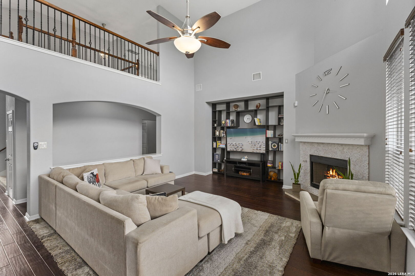Living room with double-height ceiling, beige sectional, built-in entertainment shelves, and a stone fireplace.