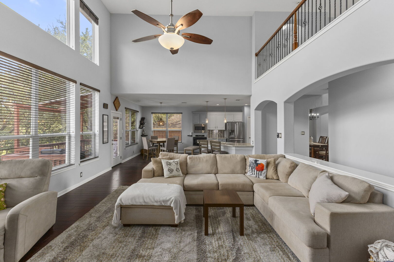 Two-story living room with beige sectional, large windows, and an open kitchen and dining area.