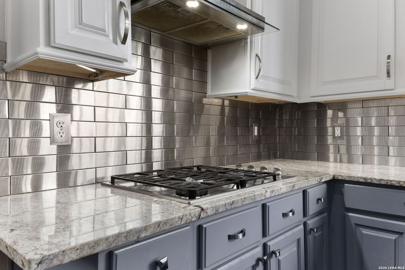 Kitchen gas cooktop beneath a stainless steel range hood, with a reflective backsplash and granite counters.