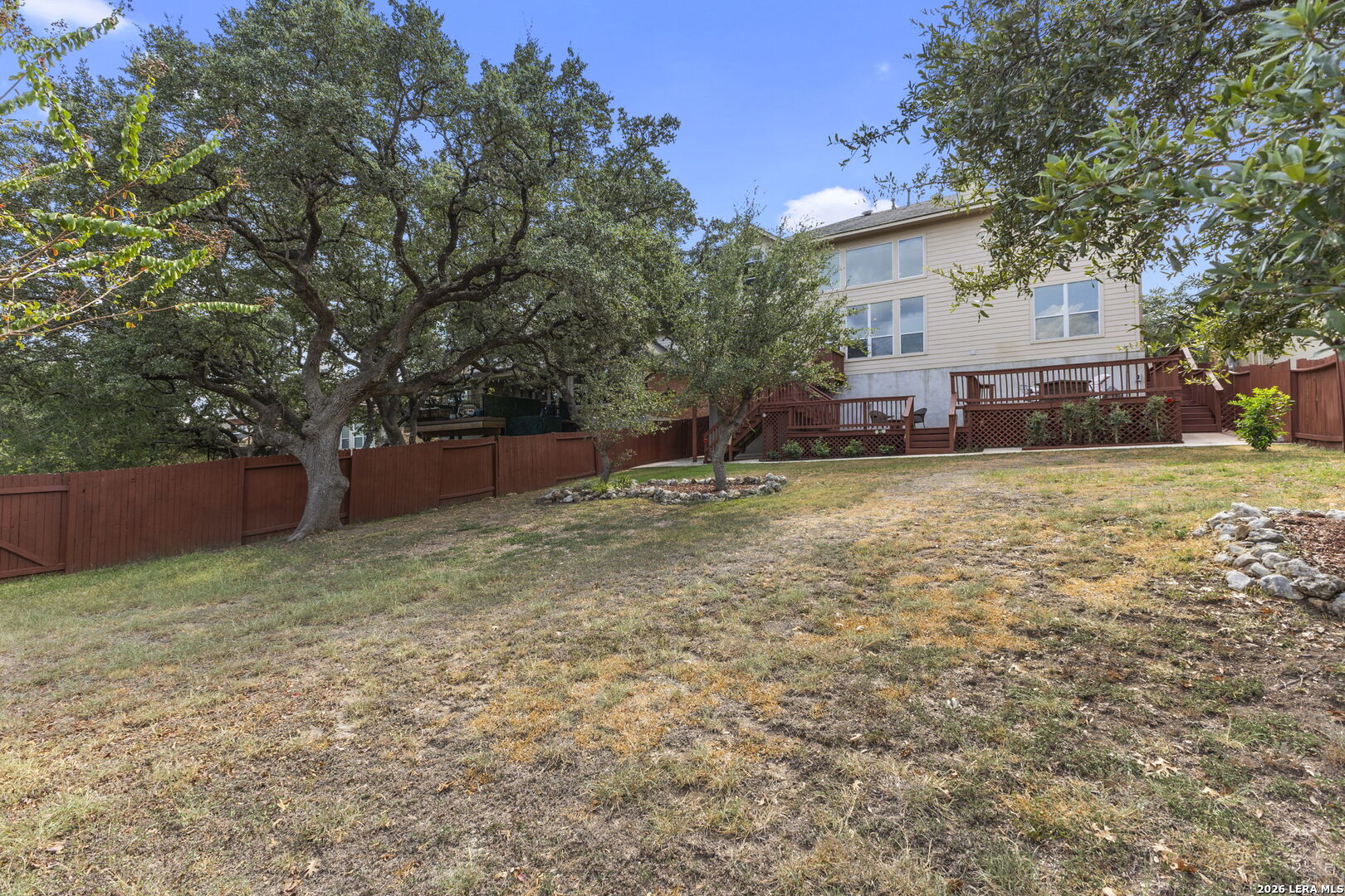 Rear of a two-story beige house with a raised deck, red fence, and mature trees.