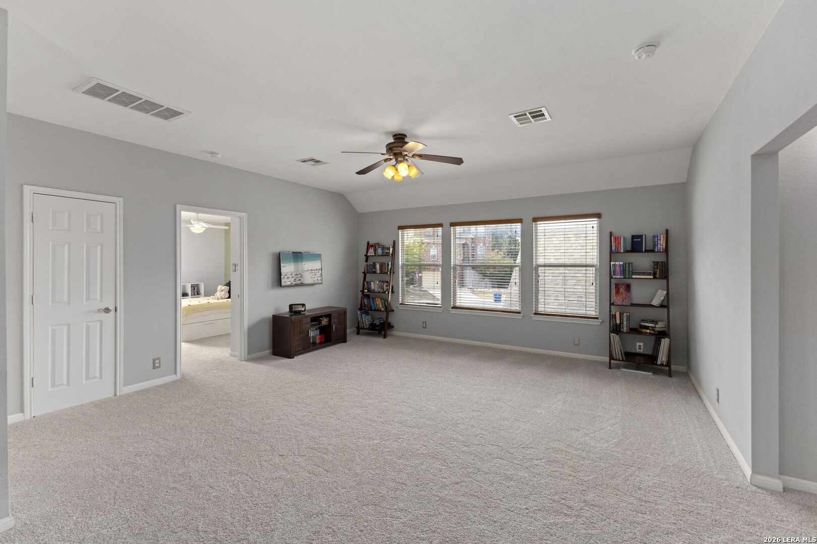 Carpeted bonus room with gray walls, three windows, bookshelves, and a ceiling fan.