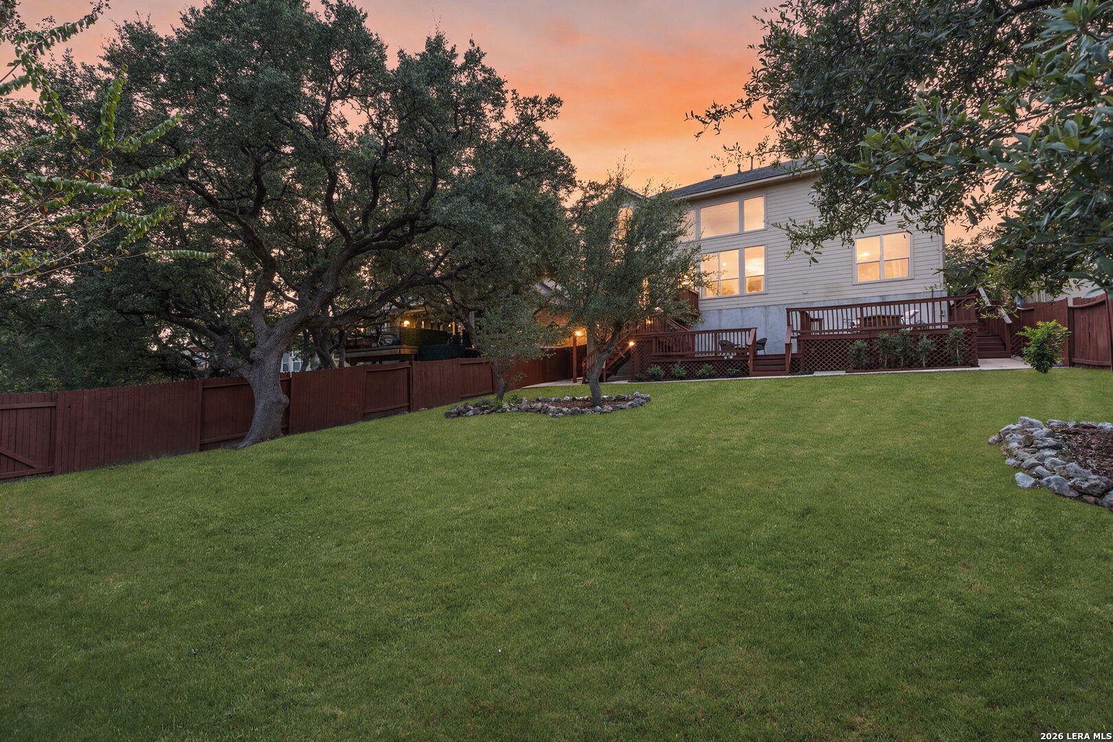Two-story home with raised deck behind a broad lawn and mature oak trees at dusk.