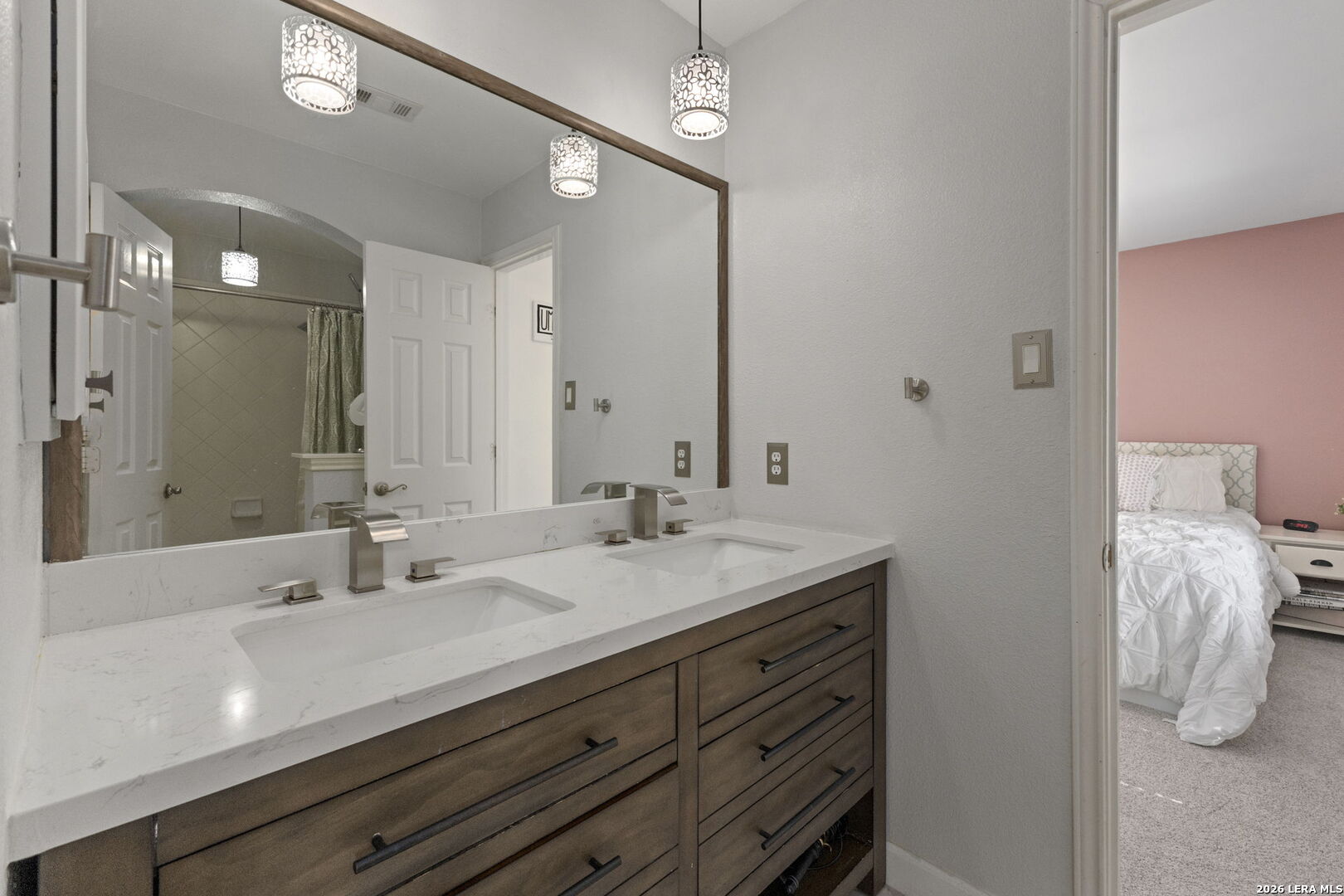 Bathroom double vanity with a white countertop and patterned pendant lights, adjoining a bedroom with a pink wall.