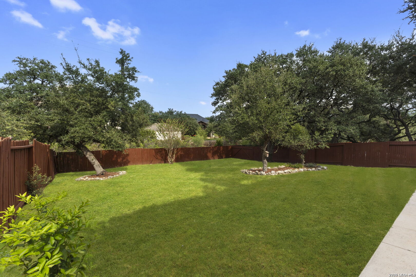 Spacious fenced backyard with a broad lawn, mature trees, and stone-edged landscaping.