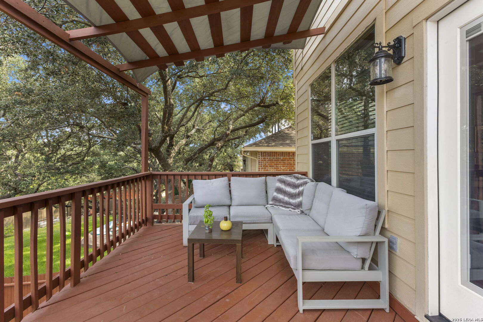 Covered wood deck with gray sectional seating beneath a pergola, overlooking leafy trees.