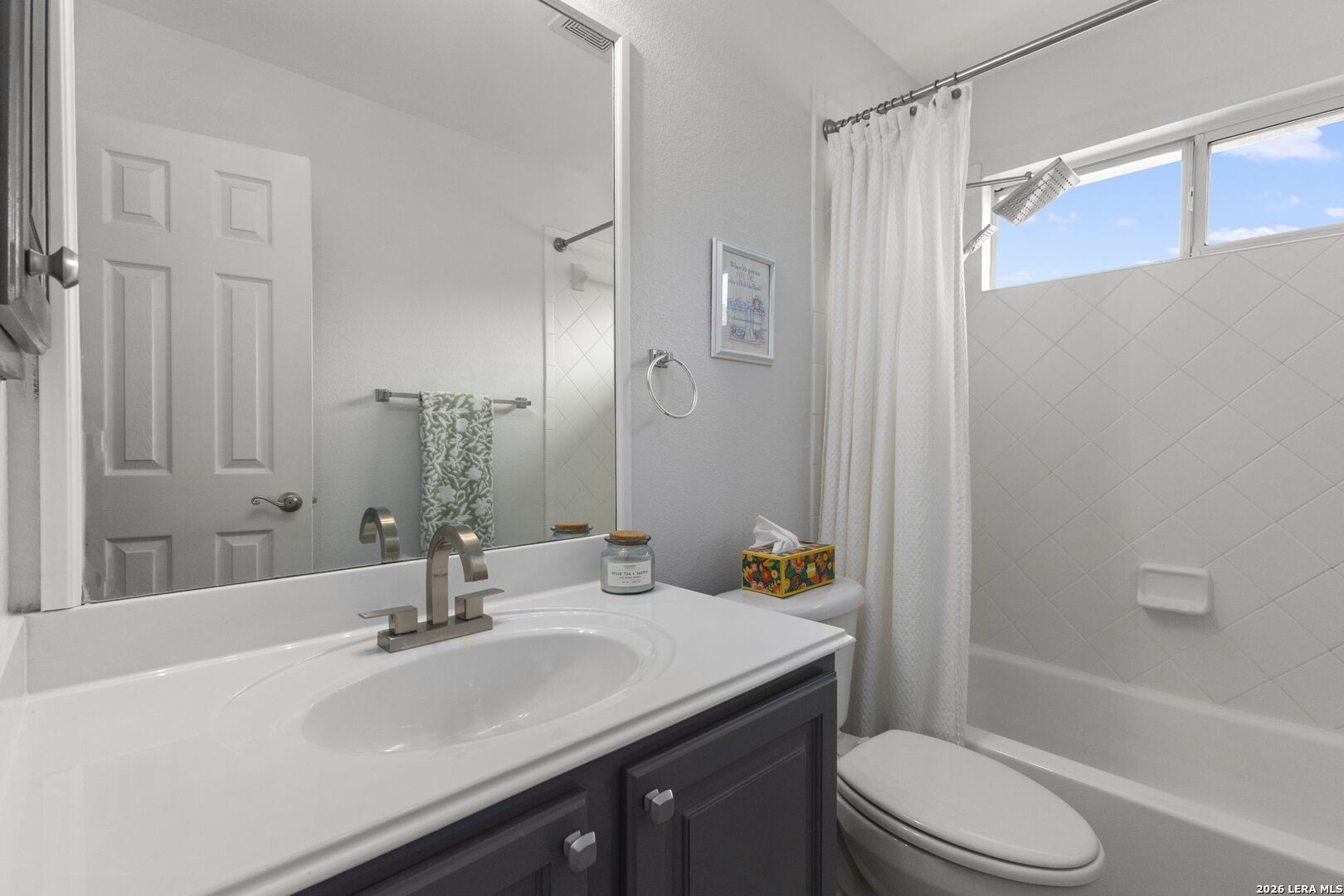Bathroom with a white vanity, large mirror, and tub beneath a high window.