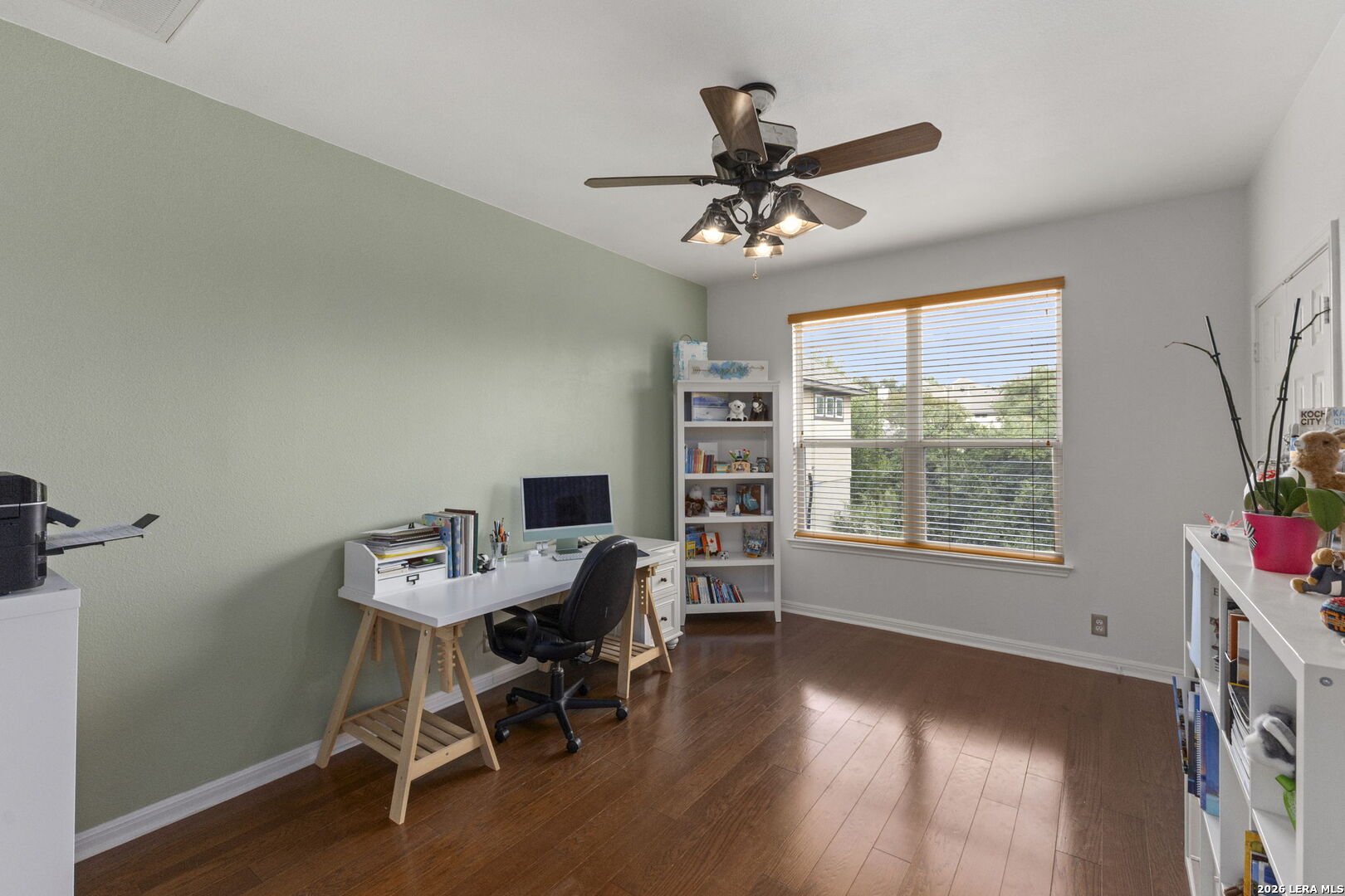Furnished home office with a white desk, tall bookcase, and large window with wooden blinds.