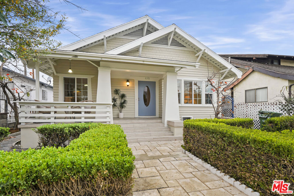 Bungalow with a broad gabled roof, covered porch, blue front door, and trimmed hedges.