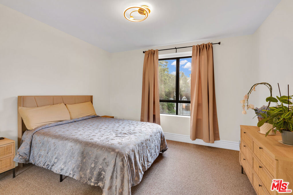 Bedroom with a patterned bedspread, tan curtains, and a light wood dresser topped with plants.