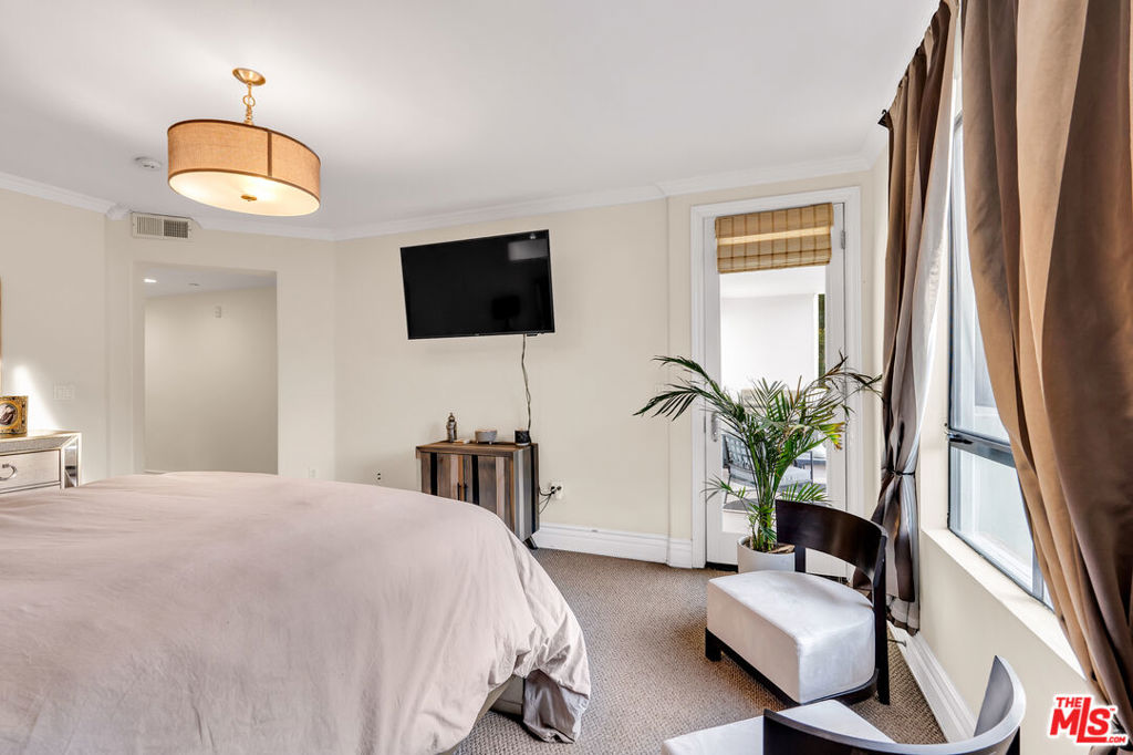 Furnished bedroom with a large bed, wall-mounted TV, potted palm, and tall curtained windows.