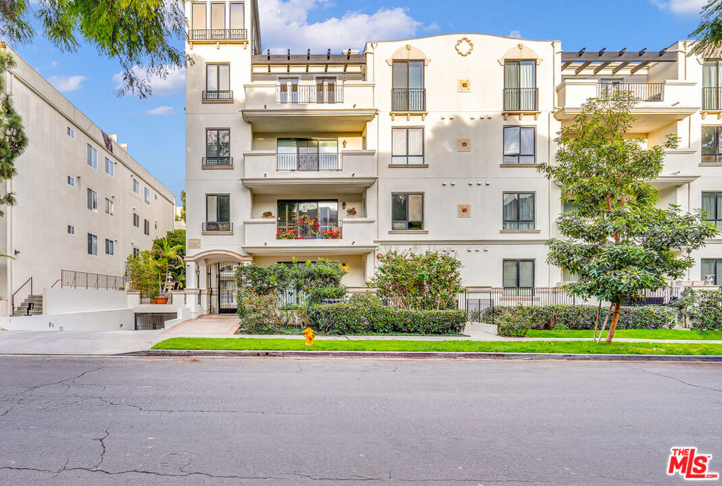 Cream-colored apartment building with balconies, leafy landscaping, and a fire hydrant along the street.