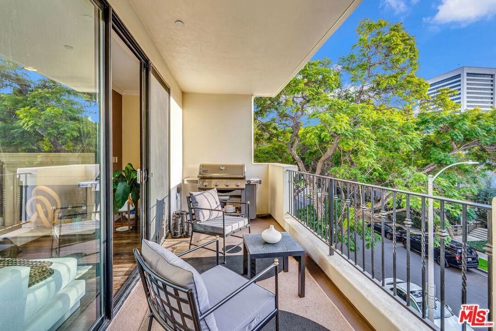Furnished apartment balcony with a grill, seating, and leafy tree views.