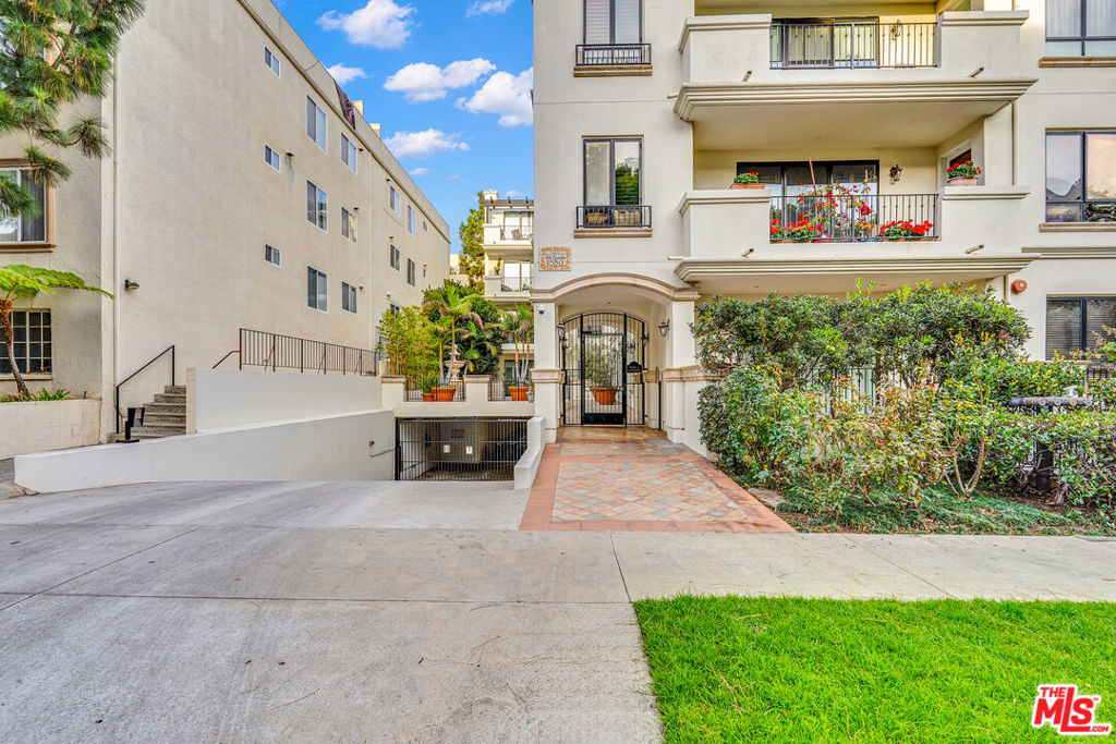 Apartment building entrance with an arched gate, tiled walkway, flowered balconies, and landscaped frontage.