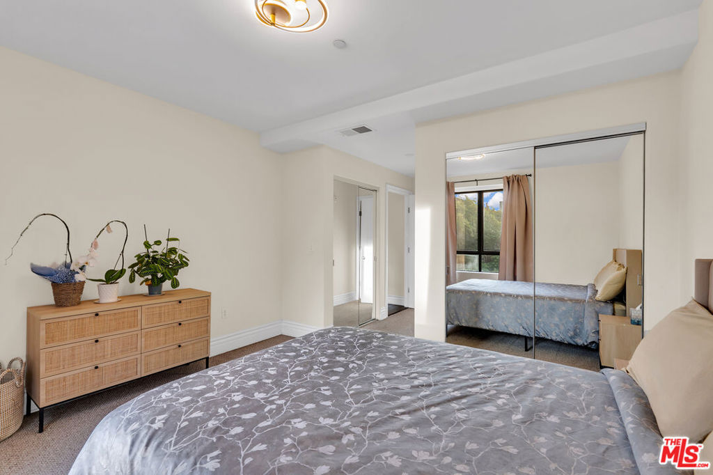 Bedroom with a gray floral bedspread, mirrored closet doors, and a light wood dresser with plants.