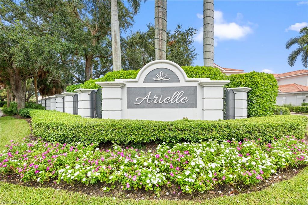 Arielle community entrance sign framed by flowering plants, hedges, and palm trees.