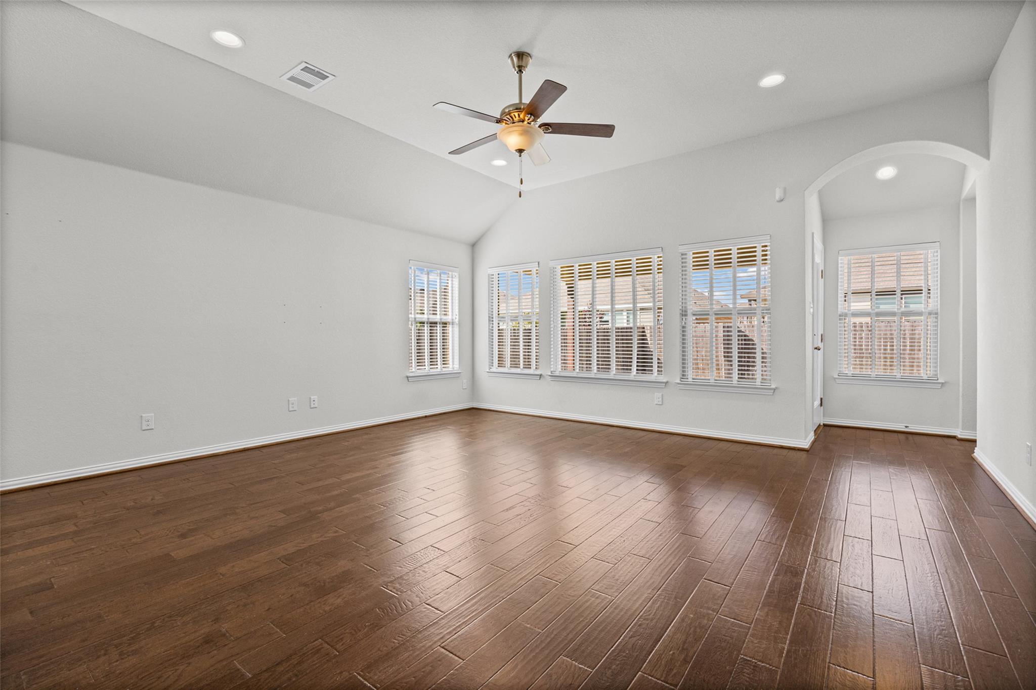 Vacant living room with dark wood floors, vaulted ceiling, ceiling fan, and multiple windows with blinds.