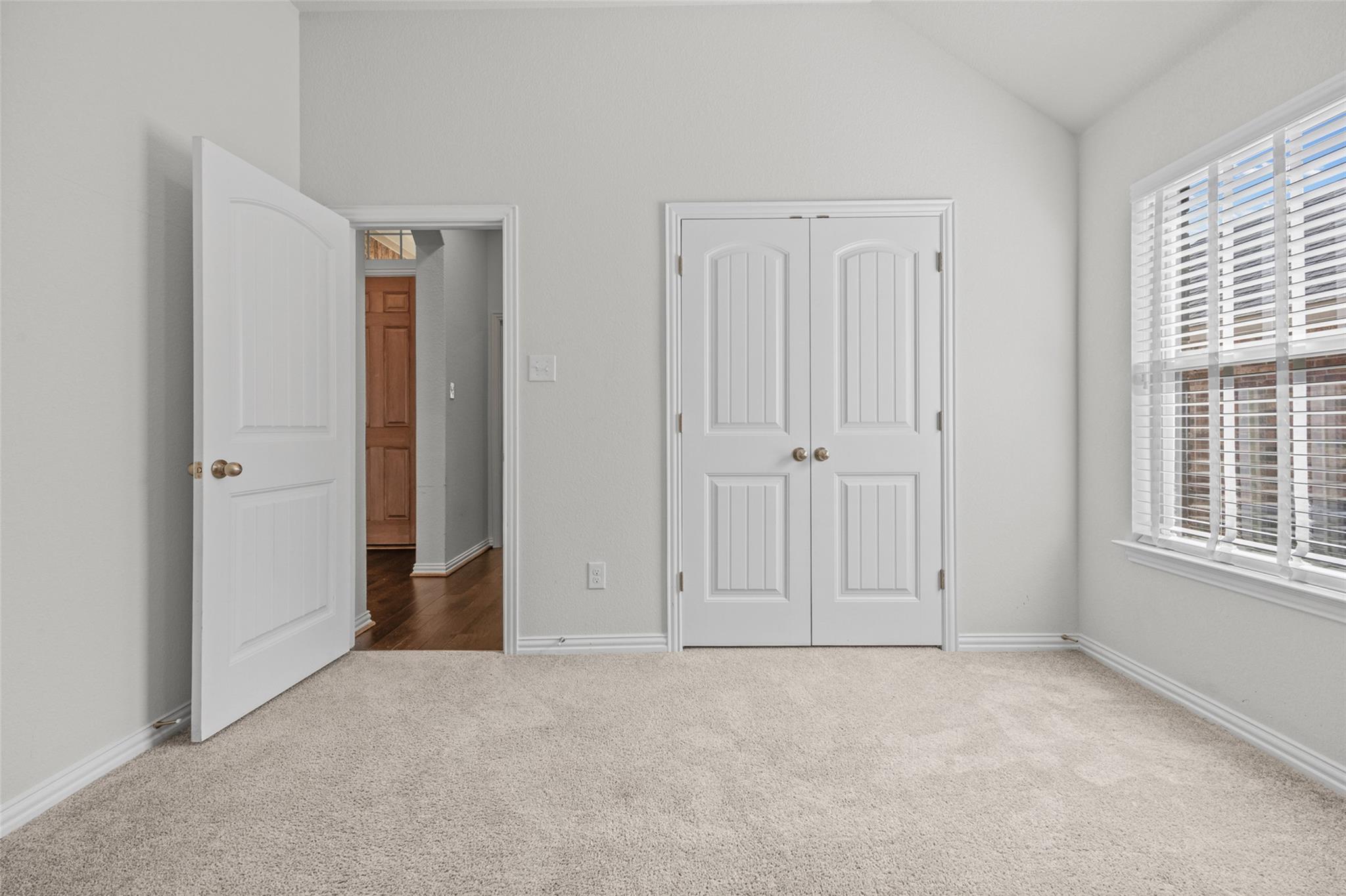 Empty carpeted bedroom with white double closet doors and a window with blinds.